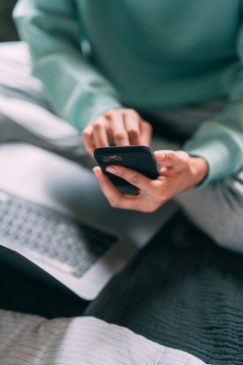 Person browsing phone on a bed next to laptop